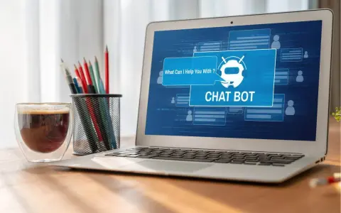 Na ekranie laptopa widać interfejs chatbota, który oferuje pomoc. Obok stoi szklanka kawy i pojemnik z przyborami do pisania.