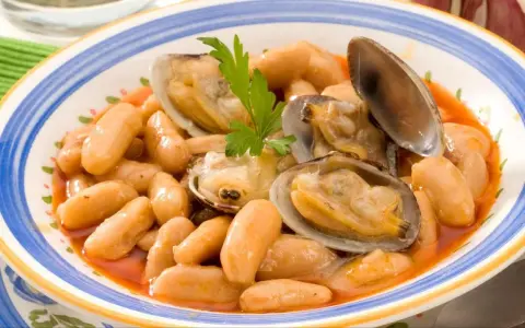 Plato de fabes con almejas, un guiso tradicional asturiano con judías blancas tiernas y jugosas almejas, decorado con perejil fresco.
