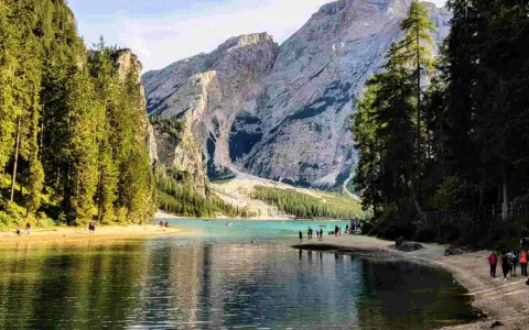 Sentiero da Valdaora al Lago di Braies: acque turchesi, montagne imponenti e pinete rigogliose. Turisti passeggiano sulla riva sabbiosa.