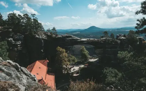 Majestatyczna kamienna brama w Czechach, otoczona zielenią i górami, z widokiem na urokliwy domek z czerwonym dachem.