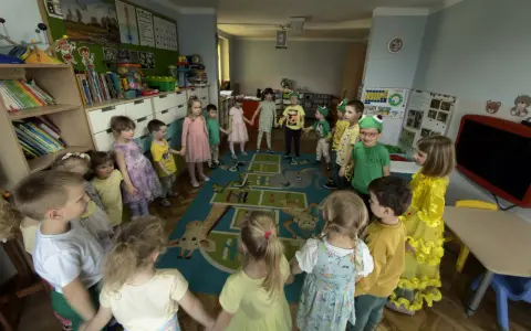 Dzieci trzymają się za ręce, tworząc krąg na dywanie z zabawnymi zwierzątkami. To radosna zabawa w **zespole szkolno przedszkolnym**.