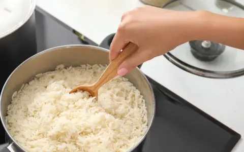 Dłoń miesza drewnianą łyżką gotujący się ryż w garnku na płycie indukcyjnej.