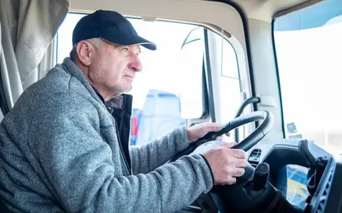 Conductor de camión con gorra y chaqueta gris, con las manos en el volante. Podría ser parte de un listado de cese de actividad de transportistas.