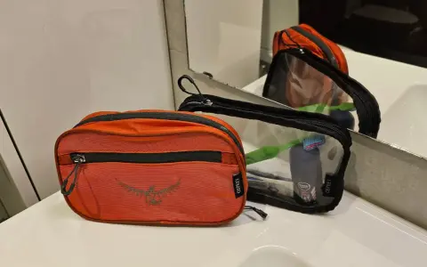 Två Osprey-necessärer, en orange och en genomskinlig, ligger på en vit bänkskiva. Den genomskinliga necessären innehåller tandborste och deodorant.