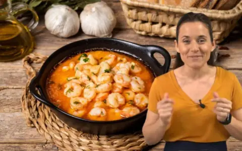 Una mujer sonríe mientras muestra cómo hacer gambas al ajillo, con un plato humeante y los ingredientes listos.