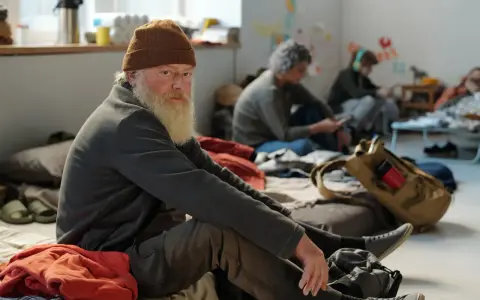 Älterer Mann mit Bart und Mütze sitzt auf dem Boden, umgeben von anderen Menschen. Eine Szene, die die Frage aufwirft: Wohin ohne Geld und Wohnung?