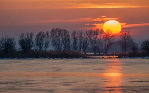 Zachód słońca nad zamarzniętym jeziorem. Piękna fotografia krajobrazowa z pomarańczowym niebem i sylwetkami drzew.