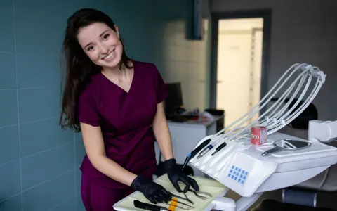 Uśmiechnięta asystentka stomatologiczna w bordowym uniformie przygotowuje narzędzia na tacce obok fotela dentystycznego.