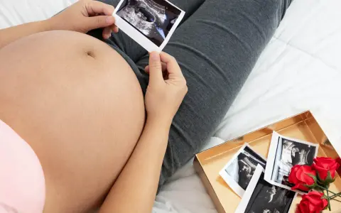 Brzuch w drugiej ciąży, trzyma zdjęcie USG. Obok pudełko z kolejnymi zdjęciami i czerwonymi różami.