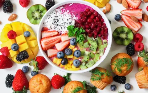Kolorowa miska smoothie z owocami, jagodami, granatem i wiórkami kokosowymi. Obok mango, truskawki, kiwi, maliny i muffinki. Idealne owoce na noc.