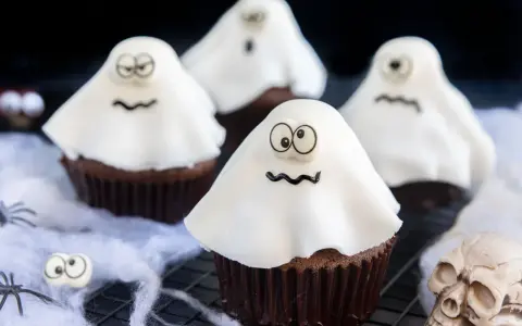 Vier schokoladige gespenster muffins mit weißen Geisterhauben und lustigen Gesichtern, dekoriert mit Spinnweben und einem Totenkopf.