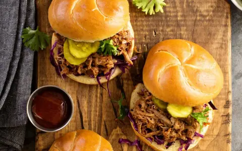 Dos sándwiches de pulled pork jugoso, con repollo morado, pepinillos y salsa barbacoa en un plato de madera.
