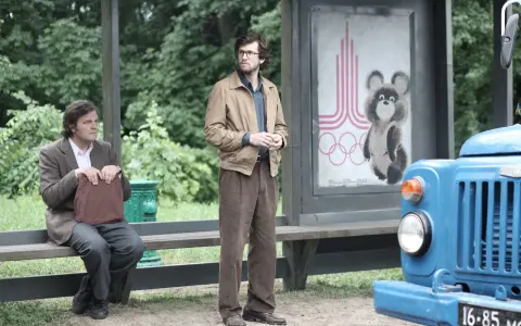 Deux hommes attendent à un arrêt de bus, un panneau des JO de Moscou 1980 avec Misha l'ours en arrière-plan. L'affaire Farewell se déroule.