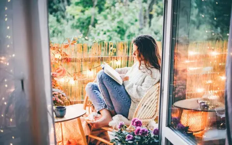 Przytulny jesienny balkon. Dziewczyna w swetrze czyta książkę, otoczona jesiennymi liśćmi i ciepłym światłem lampek.