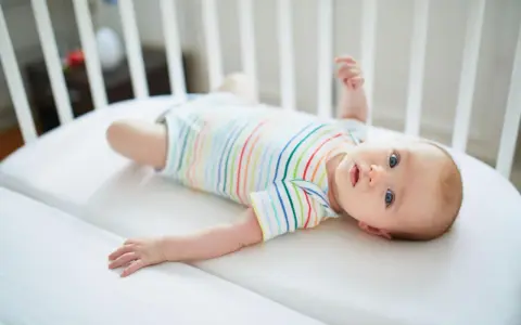 Niemowlę w kolorowym body leży w łóżeczku. Jego niebieskie oczy przypominają **perły Epsteina**, a uśmiech rozjaśnia pokój.