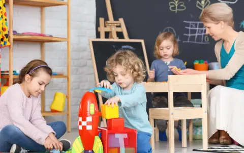 Dzieci bawią się klockami w żłobku. Nauczycielka nadzoruje zabawę, zastanawiając się, ile zarabia pomoc w żłobku.