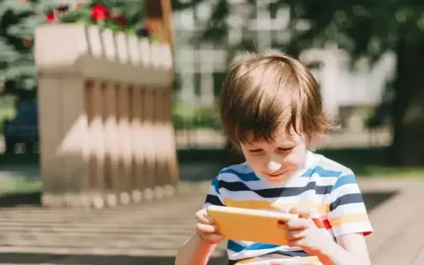 Chłopiec w kolorowym T-shircie ogląda film dla małych dzieci na żółtym telefonie, siedząc na zewnątrz.