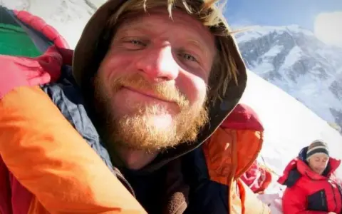 Uśmiechnięty alpinista w ciepłej kurtce, z brodą, na tle ośnieżonych gór. Może to być jego lek na chorobę wysokościową.