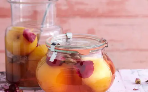 Zwei Gläser mit eingelegten Pfirsichen, verziert mit Rosenblättern und Gewürzen, auf einem rosa Holztisch.