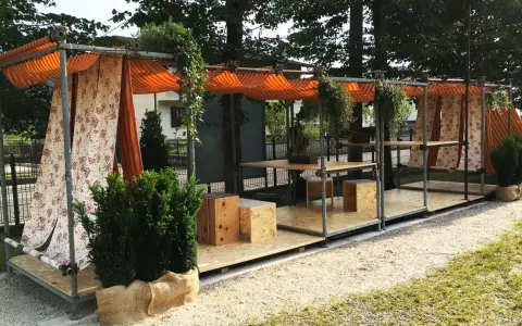 Struttura esterna con tende a righe e floreali, tavoli e sedute in legno. Un esempio di come costruire un gazebo con tubi innocenti, pratico e decorativo.