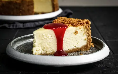 Ein Stück cremiger american cheesecake mit Keksboden, übergossen mit roter Fruchtsauce, auf einem grauen Teller.