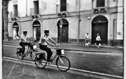 Due agenti in bicicletta pattugliano una strada italiana. Sul marciapiede, una coppia passeggia con i loro cani. Street photography Italia.