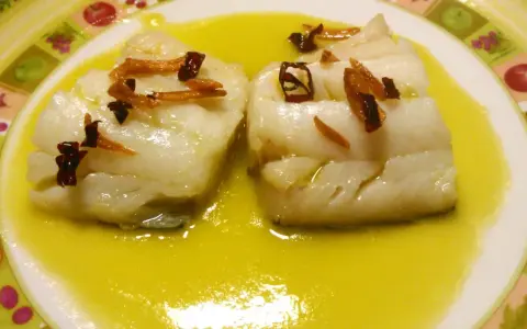 Dos trozos de bacalao al pil pil, cubiertos con ajo frito y guindilla, nadan en una salsa dorada sobre un plato decorado.