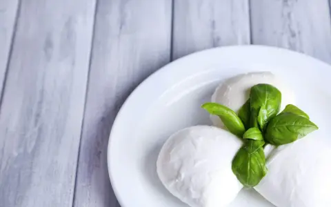 Trzy kulki serka mozzarella z listkami bazylii na białym talerzu. Idealne do sałatki, znasz ser mozzarella kcal?