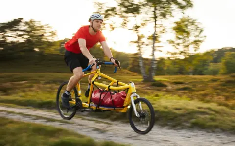 Mann fährt mit einem gelben Velo Lab Lastenrad mit roter Tasche durch eine sonnige Landschaft.