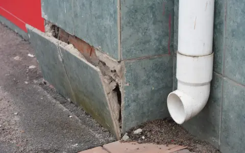 Beschädigte Sockelabdichtung am Haus. Fliesen lösen sich, darunter Ziegelmauerwerk. Ein Fallrohr leitet Wasser ab.