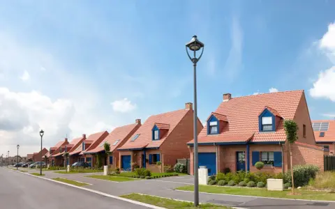 Vue d'une rue bordée de maisons de lotissement aux toits rouges et volets bleus, sous un ciel d'été.