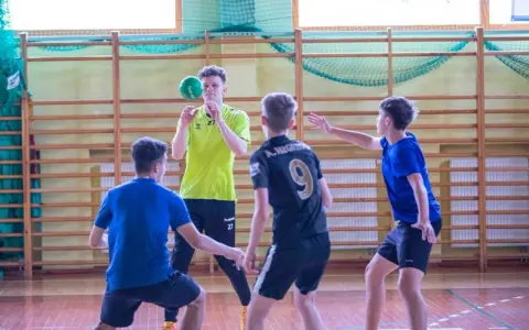 Cezary Surgiel w żółtej koszulce rzuca piłkę do ręcznej, otoczony przez kolegów z drużyny na sali gimnastycznej.
