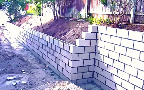 Un muro de contención de bloques de hormigón se está construyendo en un jardín, con tierra y vegetación detrás.