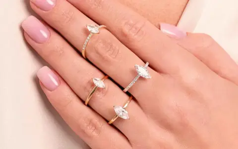 Dłonie z różowym manicure ozdobione są trzema złotymi pierścionkami z białymi kamieniami. Jeden z nich to pierścionek kwadratowy.