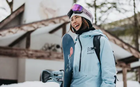 Uśmiechnięta snowboardzistka w jasnoniebieskim anoraku i goglach Anon. Gotowa na stok, trzyma deskę Burton.