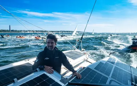 Boris Herrmann in Kiel, auf seinem Boot mit Solarpaneelen, umgeben von anderen Booten und Segelschiffen auf dem Meer.
