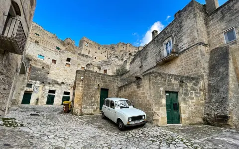 Un'auto d'epoca bianca parcheggiata tra le antiche case scavate nella roccia dei Sassi di Matera, sotto un cielo azzurro.