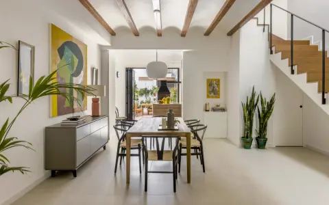 Comedor moderno en una casa entre medianeras, con vigas de madera en el techo, mesa de madera, sillas negras y una escalera de madera.