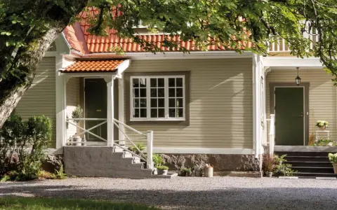 Ett charmigt hus med tegelrött tak och ljusgul fasad, redo att måla om huset. En stor ek skuggar entrén.