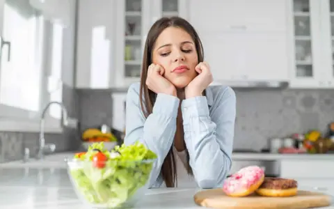 Femme triste face à une salade et des donuts, pensant aux aliments à éviter pour la dermite séborrhéique.