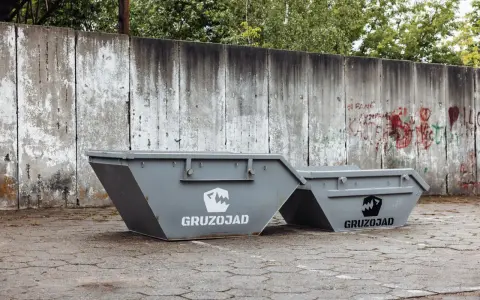 Dwa szare kontenery na śmieci zmieszane, z logo GRUZOJAD, stoją przed zniszczonym betonowym murem pokrytym graffiti.