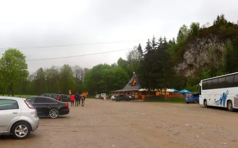 Parking przy Wąwozie Homole. Samochody, autokar i budynek zadaszony, otoczony drzewami, u podnóża skalnego zbocza.