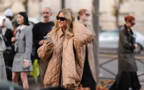 Mujer sonriente con gafas de sol y un abrigo beige claro acolchado, sosteniendo un teléfono.