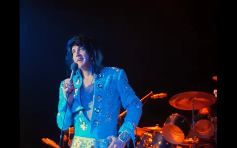 Elvis Presley, in einem glitzernden blauen Anzug, singt ins Mikrofon. Ein Bild, das an seine legendären Elvis Presley Filme erinnert.