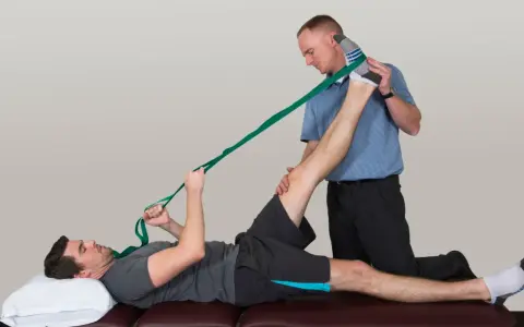 Physiotherapeut hilft Mann bei der Dehnung des Beins mit einem Band. Sanfte Gymnastik bei Bandscheibenvorfall zur Linderung.