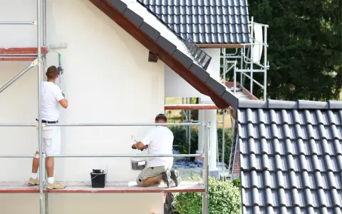 Dwóch malarzy na rusztowaniu odnawia elewację domu. Sprawdź, jaka jest malowanie elewacji cena.