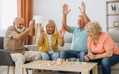 Radosna terapia zajęciowa dla seniorów: czworo uśmiechniętych osób gra w Jengę, unosząc ręce w geście zwycięstwa lub ekscytacji.