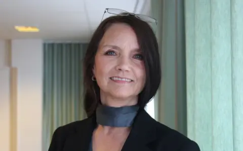 En kvinna med glasögon på huvudet ler. Hon bär en svart kavaj och en grå scarf.