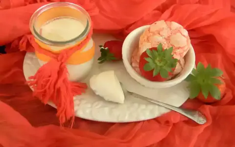 Yogur cremoso en frasco con fresas y trozos de yogur en un plato blanco sobre tela naranja.