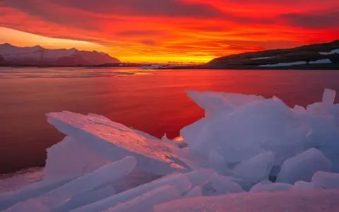 Isen glittrar i förgrunden medan himlen tycks måla solnedgång i dramatiska röda och orange toner över ett lugnt vatten.